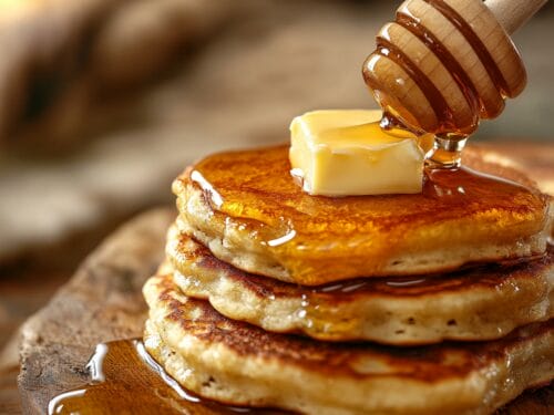 Three stacked golden pancakes with melting butter and drizzled honey on wooden board