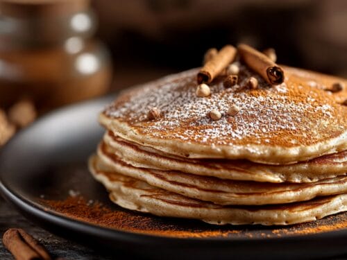 Stack of four pancakes dusted with powdered sugar and cinnamon, topped with cinnamon sticks and cardamom pods