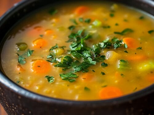 Golden soup in dark bowl topped with fresh green parsley and diced carrots
