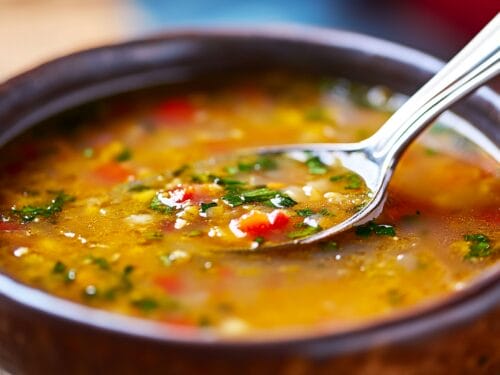 Golden broth in dark bowl with spoon, garnished with red pepper and green herbs