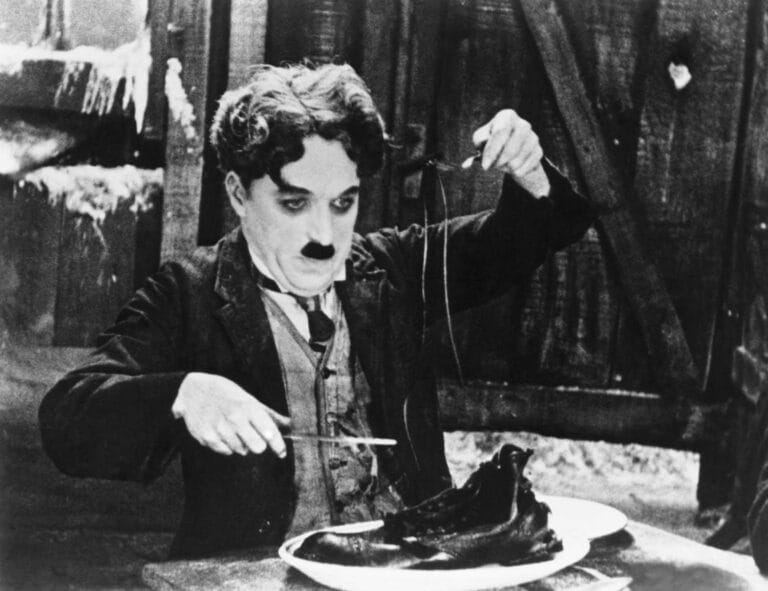 Black-and-white film still showing a man in suit and hat eating from a plate with fork and knife