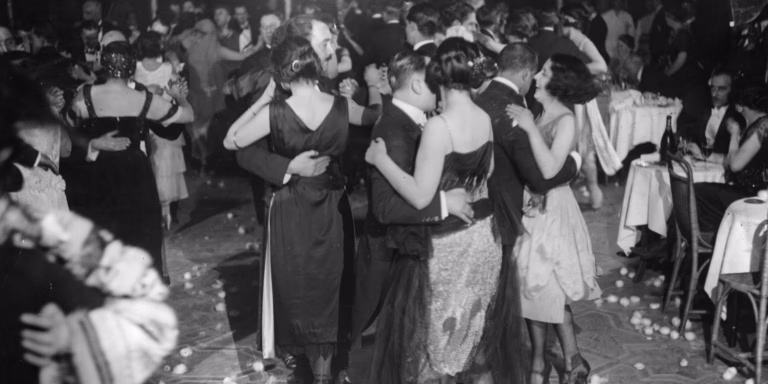 Black and white photograph of couples dancing closely together at crowded indoor party with bottles and glasses visible
