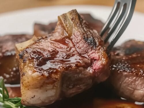 Cooked meat chops with bone on white plate, fork lifting one piece, garnished with fresh herbs