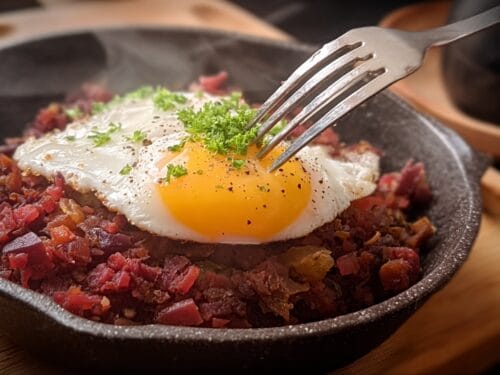 Skillet of diced red and brown meat topped with fried egg, fork poised above, garnished with parsley