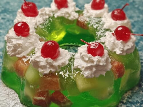 Green gelatin ring mold topped with whipped cream dollops, maraschino cherries, shredded coconut, and embedded fruit pieces
