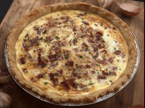 Baked savory pie in aluminum tin with bacon pieces and chives scattered across custard filling