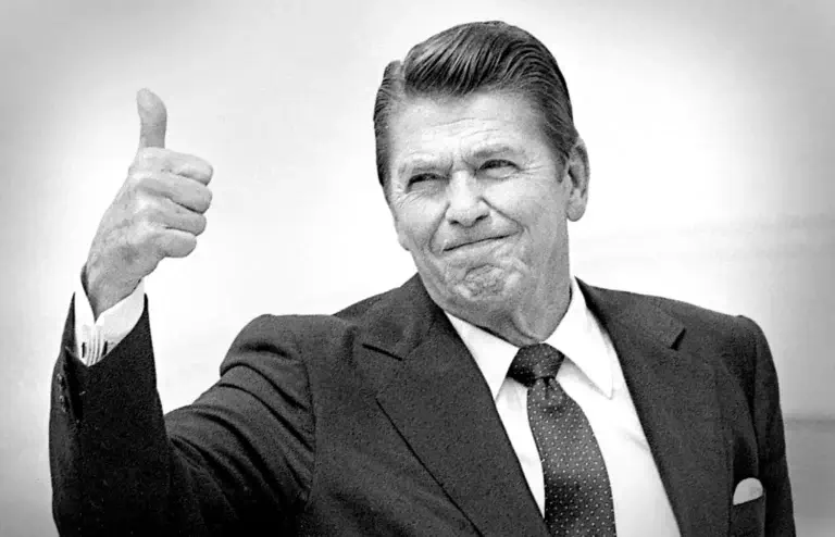 Black and white portrait of a man in dark suit and patterned tie giving a thumbs up