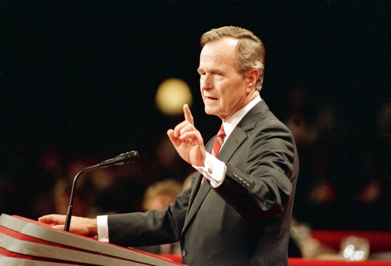 Man in gray suit and red tie speaking at podium with microphone before audience