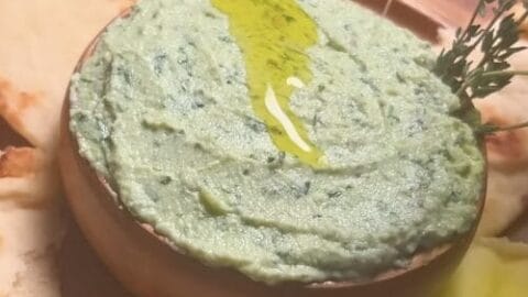 Pale green herb and cheese spread in a shallow bowl, drizzled with olive oil and garnished with fresh herbs