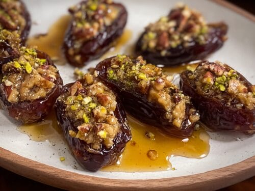 Halved dark dates filled with chopped nuts and green pistachios, seated in pooled golden oil on ceramic plate