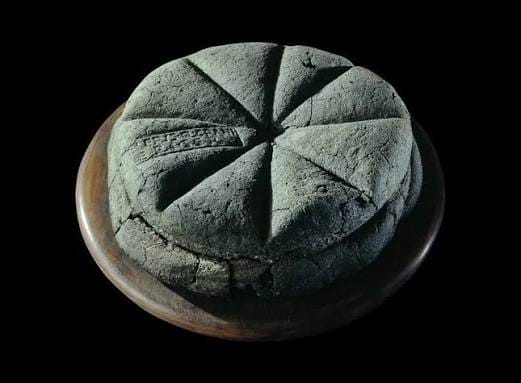 Round loaf scored into eight wedge-shaped sections, resting on a clay plate against black background