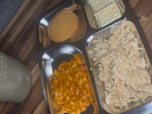 Overhead view of military ration components in metal trays: tan sauce, orange beans, shredded meat, crackers, and a container of spread