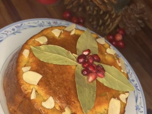 Round cake topped with sliced almonds, bay leaves, and red berries on blue-and-white plate