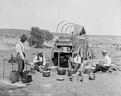 Black and white photograph of people preparing food beside a covered wagon in an arid landscape