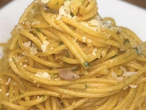 Heap of spaghetti coated in pale sauce with grated cheese, green herb, and black pepper on white plate