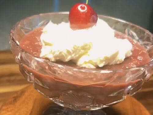 Stemmed glass bowl with brown pudding, white cream dollop, yellow shavings, and cherry with stem on top