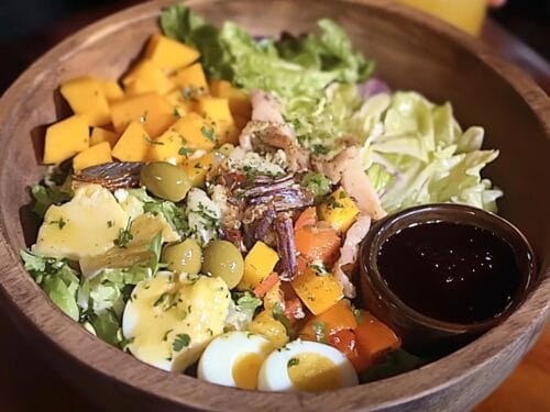 Wooden bowl with lettuce, shredded meat, potatoes, yellow cheese, red peppers, olives, egg halves, and dark dipping sauce