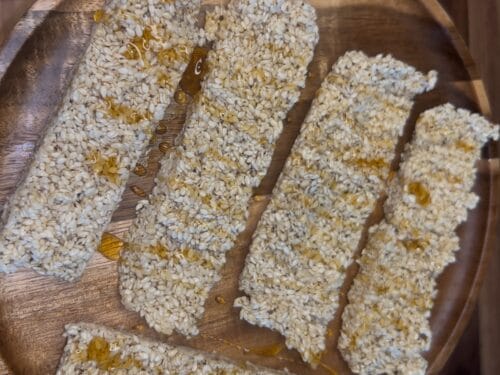Five rectangular bars coated in sesame seeds with honey drizzle, arranged on a wooden tray