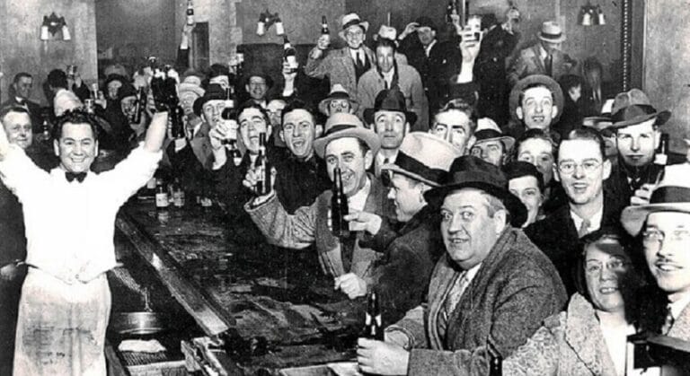 Black and white photograph of a crowded indoor gathering with men in suits and hats holding bottles and glasses