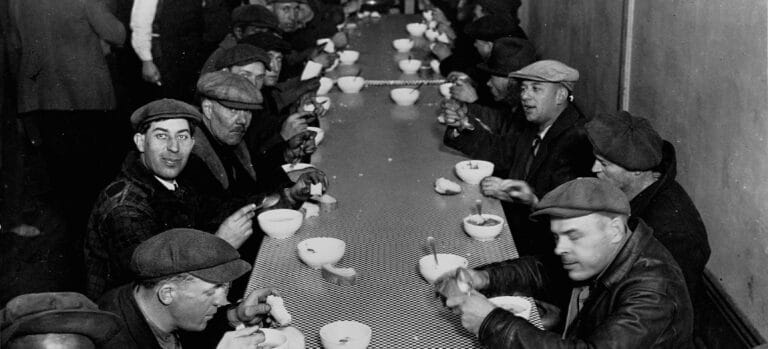 Men in flat caps seated at long table eating soup from white bowls with spoons
