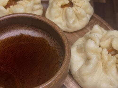 Pale cream-colored steamed buns with gathered tops arranged on wooden board beside dark liquid in wooden bowl