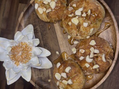 Round golden-brown cakes topped with sliced almonds on a wooden plate, beside a white lotus flower