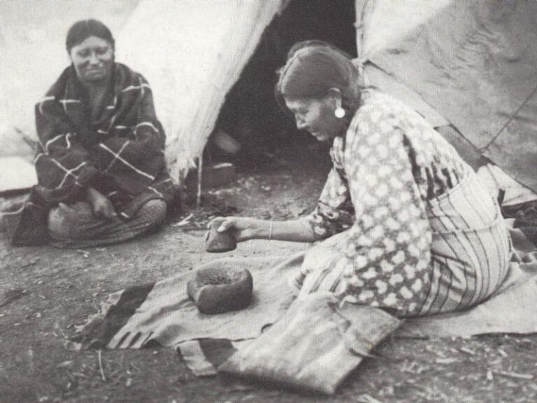 Black and white photograph showing two people inside a tent preparing food on the ground.