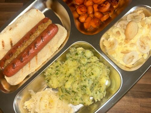 Four metal trays holding a hot dog on bun, glazed carrots, mashed potatoes with herbs, and shredded potatoes