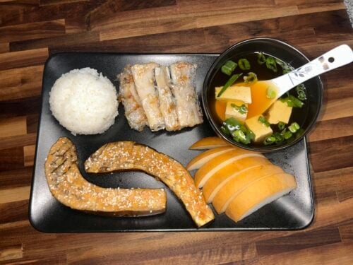 Black plate with white rice, sliced grilled meat, sesame-crusted eel, sliced bread, and bowl of tofu soup with green peppers