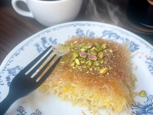 Golden shredded pastry dessert topped with pistachios and spices on blue-patterned plate with fork