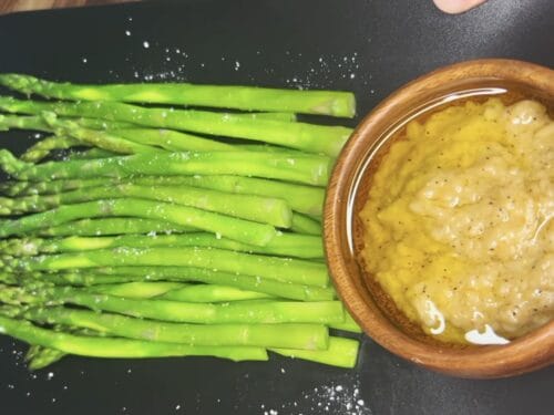 Cooked asparagus spears on black plate with yellow mustard sauce in wooden bowl