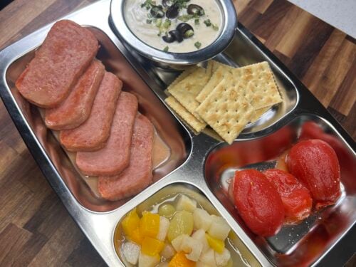 Sliced salami, crackers, cream dip with jalapeños, canned tomatoes, and mixed pickled vegetables on a divided tray