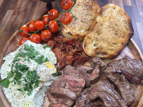 Wooden board with seared beef medallions, grilled tomatoes, toasted bread, creamed cheese with herbs, and dark wine