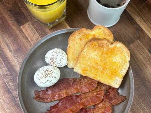 Plate with bacon strips, poached eggs with pepper, and buttered toast beside orange juice and black coffee