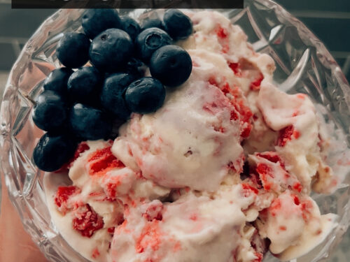 Creamy white ice cream with red fruit pieces and fresh blueberries in a glass bowl