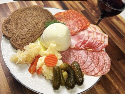 White plate with sliced salami, whole onion, pickles, potato salad, sliced bread, and dark beverage
