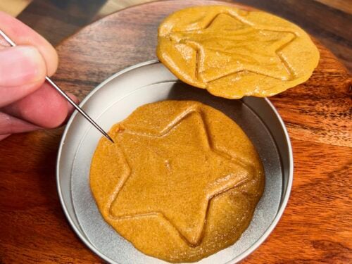 Two tan whipped candies with drawn outlines, one in white bowl, one held by fingers above wooden surface