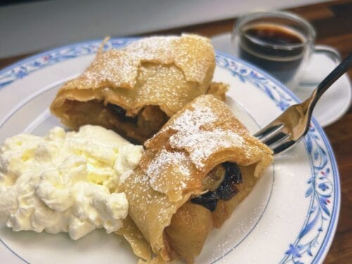 Rolled pastry dusted with powdered sugar, filled with dark fruit, served with whipped cream and coffee
