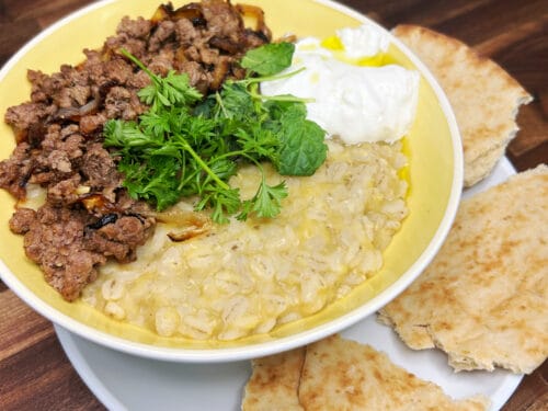 Yellow bowl of pale grain porridge topped with browned meat, parsley, white yogurt, olive oil, and flatbread on the side