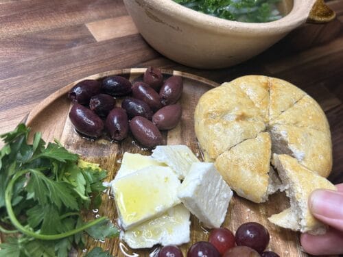 Ceramic bowl with broth and greens, wooden board with olives, grapes, cheese, and sliced bread