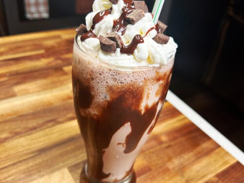 Milkshake in tall glass with whipped cream, chocolate syrup drizzle, chocolate chunks, and striped straw on wooden table