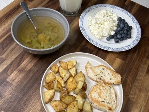 Four dishes on wood surface: soup with spoon, glass of milk, cottage cheese with blueberries, roasted potato wedges and seasoned fish fillets