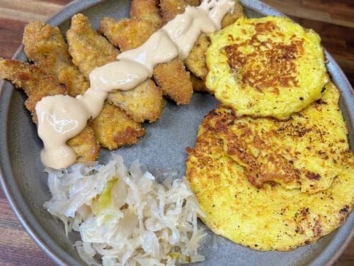 Gray plate with breaded cutlets, cream sauce, yellow pancakes, and shredded cabbage on wooden surface
