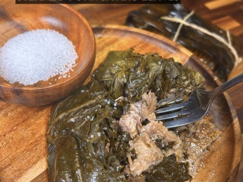 Unwrapped bundle of dark green taro leaves revealing shredded pork inside, fork nearby, wooden bowl of sea salt at left