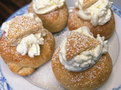 Four round pastry buns with powdered sugar and cream filling on decorative blue-and-white plate