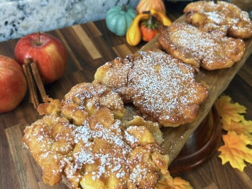 Powdered sugar-coated fried pastries on wooden board with red apples, decorative gourds, and cinnamon sticks