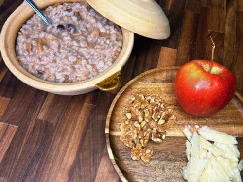 Wooden bowl of cooked grain porridge with lid, wooden board holding red apple, walnuts, and sliced cheese