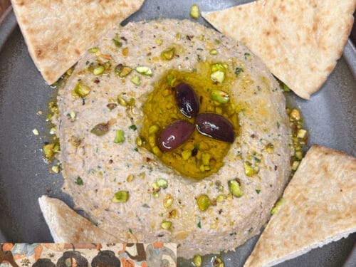 Beige puree topped with olive oil, pistachios, and two dark olives, surrounded by pita chips