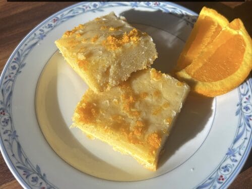 Two thick squares of yellow cake on a decorative blue-and-white plate, topped with powdered sugar and accompanied by a fresh orange wedge