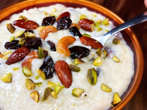 White rice pudding in orange-rimmed bowl garnished with almonds, raisins, cashews, and pistachios